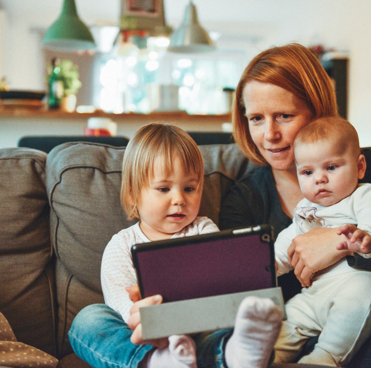 A Mother with her two children holding an tablet device , one is a toddler and one is a baby,