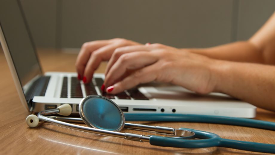 Hands typing on a laptop on a desk with a stethoscope in the foreground